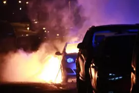 Brennendes Auto auf einer Straße bei Nacht, davor ein parkendes Fahrzeug.