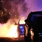 Brennendes Auto auf einer Straße bei Nacht, davor ein parkendes Fahrzeug.