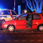 Rotes Auto mit geöffneten Türen und Kofferraum steht nachts auf Straße, im Hintergrund Polizeiwagen mit Blaulicht und zwei Personen.