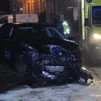 Schwarzes Auto mit stark beschädigter Front steht nachts am Straßenrand, daneben ein gelbes Einsatzfahrzeug mit Blaulicht.