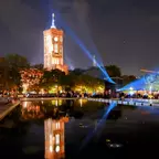 Das beleuchtete Rote Rathaus in Berlin spiegelt sich nachts im Wasser der davorliegenden Wasserkaskaden, umgeben von Bäumen und Menschen.