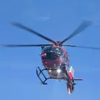 Hubschrauber mit laufenden Rotorblättern fliegt vor klarem Himmel, Scheinwerfer leuchtet nach unten.