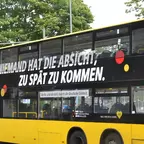 Gelber Doppeldeckerbus mit schwarzer Werbefläche und weißer Aufschrift 'NIEMAND HAT DIE ABSICHT, ZU SPÄT ZU KOMMEN.'