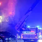 Ausgefahrene Drehleiter der Feuerwehr mit Blaulicht, daneben mehrere geparkte Autos und Feuerwerk am Nachthimmel