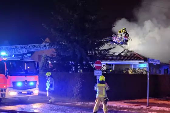 Feuerwehrleute mit gelben Helmen und Schutzkleidung löschen mit Drehleiter einen Dachstuhlbrand an einem Wohnhaus bei Nacht, Rauch steigt auf
