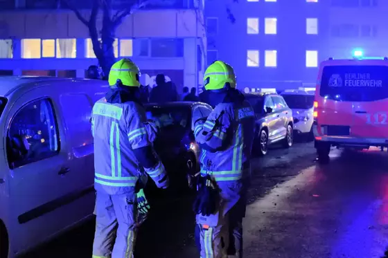 Zwei Feuerwehrleute in Schutzkleidung und gelben Helmen stehen bei geparkten Autos auf einer Straße, im Hintergrund ein Einsatzfahrzeug der Berliner Feuerwehr mit Blaulicht