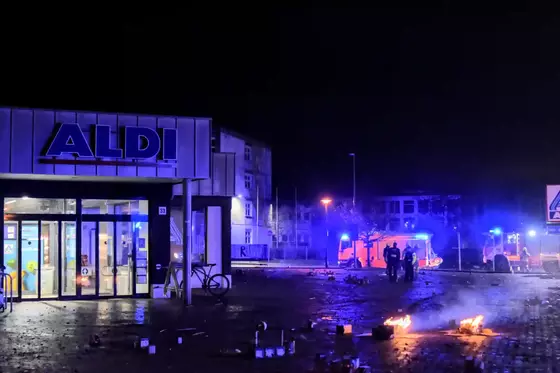 Brennende Feuerwerksbatterien liegen auf dem Parkplatz vor einem ALDI-Supermarkt, im Hintergrund sind Feuerwehrfahrzeuge mit Blaulicht und Einsatzkräfte zu sehen