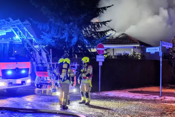 Drei Feuerwehrleute in Schutzkleidung und Atemschutz stehen vor einem brennenden Hausdach, im Hintergrund eine Drehleiter und Rauchentwicklung