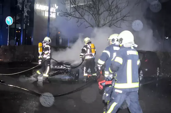 Feuerwehrleute in Schutzkleidung und Helmen führen nachts Löscharbeiten an zwei brennenden Fahrzeugen durch, umgeben von Rauch