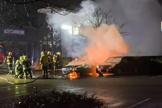 Feuerwehrleute löschen nachts zwei brennende Fahrzeuge auf einer Straße, umgeben von Rauch und Flammen