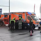 Unfallstelle mit einem verunfallten dunklen PKW, der mit einem Rettungswagen kollidiert ist, umgeben von mehreren Einsatzkräften in Schutzkleidung und Verkehrskegeln auf nasser Straße.