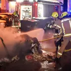 Feuerwehrleute löschen nachts einen brennenden Abfallcontainer auf einer Straße vor einem Feuerwehrfahrzeug
