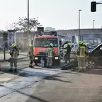 Feuerwehrleute in Schutzkleidung stehen vor einem Einsatzfahrzeug auf einer Straße neben einem beschädigtem schwarzen Auto.