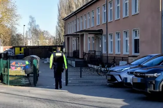 Ein Polizist in schwarzer Uniform mit gelben Reflexstreifen geht auf einem Gehweg neben einem mehrstöckigen Wohngebäude mit mehreren geparkten Autos.