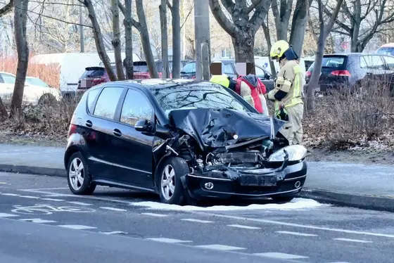 Schwarzes Auto mit stark beschädigter Frontpartie steht am Straßenrand, daneben zwei Personen in Warnkleidung und Helm