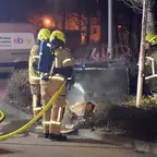 Drei Feuerwehrleute in Schutzkleidung und Helmen löschen mit einem Schlauch einen kleinen Brand an einem Straßenrand bei Nacht