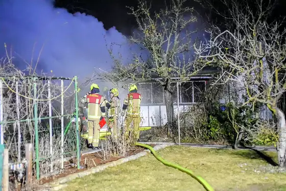 Drei Feuerwehrleute in Schutzkleidung stehen vor einem brennenden Gebäude, aus dem Rauch aufsteigt, im Garten mit Bäumen und einem Zaun