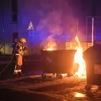 Feuerwehrmann in Schutzkleidung löscht brennenden Müllcontainer bei Nacht mit Löschschlauch