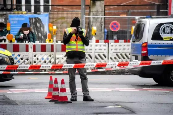 Polizist mit Warnweste und Mütze fotografiert eine Hülse auf dem Boden hinter Absperrband an einem Tatort im Straßenbereich