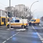 Zwei gelbe Einsatzfahrzeuge mit Blaulicht stehen an einer Kreuzung neben einer gelben Straßenbahn und mehreren Autos auf einer nassen Straße in einer städtischen Umgebung mit Hochhäusern im Hintergrund