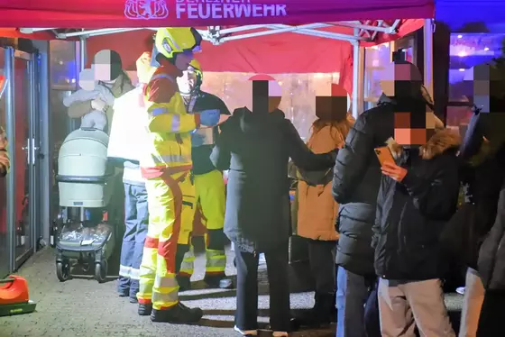 Mehrere Personen stehen bei einer Sammelstelle zur Sichtung von Verletzten unter einem Zelt mit der Aufschrift "FEUERWEHR", darunter zwei Einsatzkräfte in gelb-roter Schutzkleidung und Helm.