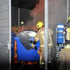 Zwei Feuerwehrleute in Schutzkleidung und Helmen löschen einen brennenden blauen Müllcontainer hinter einem Metallzaun