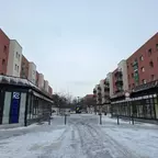 Schneebedeckte Fußgängerzone mit modernen Wohngebäuden und Ladenlokalen an der Hellersdorfer Promenade unter bewölktem Himmel