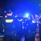 Mehrere Polizisten mit reflektierenden "POLIZEI"-Schriftzügen auf der Kleidung stehen nachts auf einer Straße mit geparkten Fahrzeugen und eingeschaltetem Blaulicht.