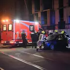 Feuerwehrleute und Polizisten stehen bei einem Unfall an einem beschädigten Auto neben einem Rettungswagen mit eingeschaltetem Blaulicht in der Nacht