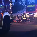Verletzter sitzt auf dem Boden zwischen zwei Feuerwehrfahrzeugen, Feuerwehrleute in Schutzkleidung versorgen ihn mit medizinischem Material