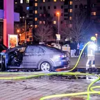 Feuerwehrmann mit Helm löscht nachts mit Schlauch einen brennenden Pkw mit geöffneter Motorhaube auf einem Parkplatz vor Wohngebäuden