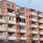 Mehrstöckiges Wohngebäude mit Balkonen und teils verputzter Fassade in einem urbanen Umfeld. Ein Balkon hat deutliche Brandspuren.