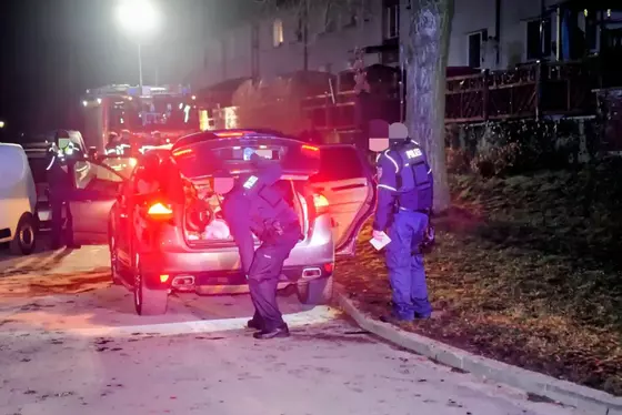 Zwei Polizisten durchsuchen nachts den geöffneten Kofferraum eines Autos auf einer Straße vor Wohnhäusern.