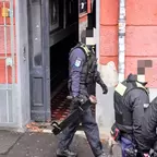 Mehrere Polizisten in Uniform stehen vor einem roten Gebäude mit offenem Eingang, einer trägt eine Ramme in der Hand.
