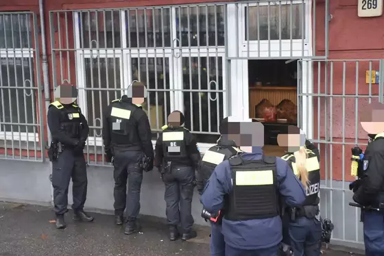 Mehrere Polizeibeamte in Uniform stehen vor einem geöffneten Tor eines Gebäudes, durch das ein Innenraum mit Möbeln sichtbar ist.