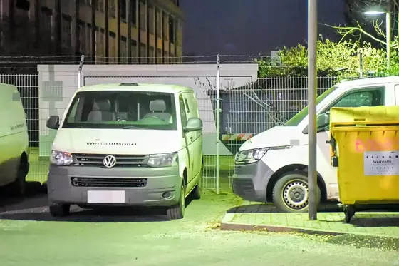 Zwei weiße Transporter vor einem beschädigten Metallzaun mit Stacheldraht an einem beleuchteten Parkplatz bei Nacht