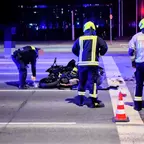 Polizeibeamter und zwei Feuerwehrleute in Schutzkleidung untersuchen nachts eine Unfallstelle mit einem umgestürzten Motorrad auf einer Kreuzung, ein Verkehrshütchen steht auf der Straße.