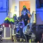 Drei Personen werden von Feuerwehrleuten in Schutzkleidung aus einem Haus mit beleuchtetem Eingang begleitet.
