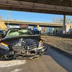Schwarzes Mercedes-Auto mit stark beschädigter Frontpartie steht auf einer Straße unter einer Brücke, im Hintergrund Einsatzfahrzeuge und Personen