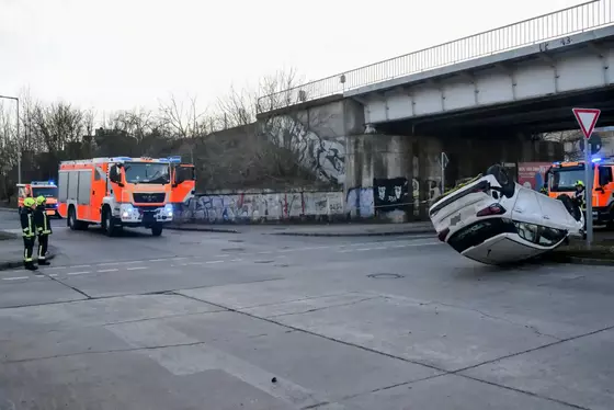Ein auf der Seite liegendes Auto auf einer Kreuzung unter einer Brücke mit zwei orangefarbenen Einsatzfahrzeugen und zwei Einsatzkräften in Warnkleidung.