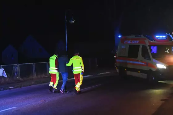 Zwei Rettungsdienstmitarbeiter in gelb-roter Schutzkleidung begleiten eine Person zu einem Rettungswagen mit Blaulicht auf einer dunklen Straße