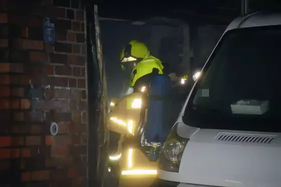 Feuerwehrkräfte mit Atemschutzgeräten und reflektierender Schutzkleidung stehen neben einem weißen Fahrzeug vor einer Backsteinwand.