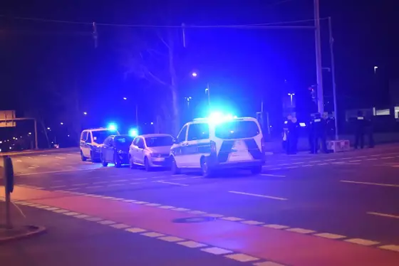 Mehrere Fahrzeuge mit eingeschaltetem Blaulicht stehen nachts auf einer Straße, daneben stehen mehrere Personen in dunkler Kleidung