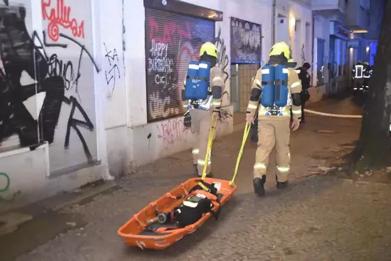 Zwei Feuerwehrleute in Schutzkleidung ziehen eine orangefarbene Trage mit einer Person durch eine mit Graffiti bedeckte Straße.