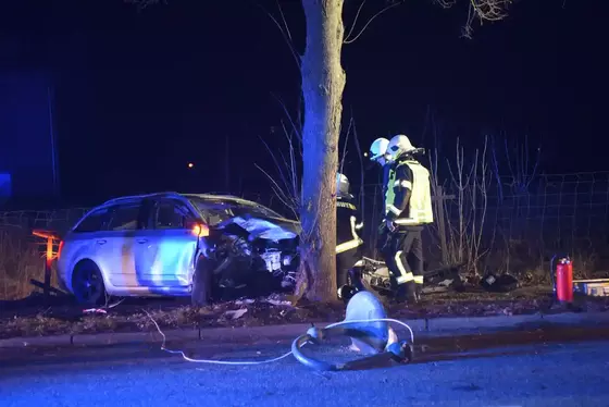 PKW mit stark beschädigter Front steht an einem Baum, Feuerwehrleute in Schutzkleidung arbeiten an der Unfallstelle bei Nacht