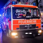 Feuerwehrfahrzeug der Marke MAN mit eingeschaltetem Scheinwerfer steht nachts in einer Straße mit weiteren Einsatzfahrzeugen im Hintergrund.