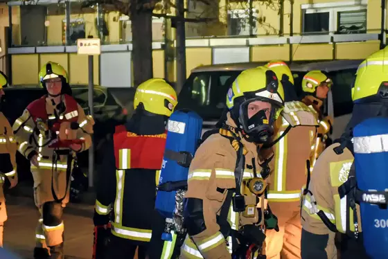 Mehrere Feuerwehrleute in Schutzkleidung und Helmen mit Atemschutzgeräten stehen nachts auf einer Straße vor geparkten Autos.