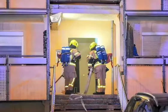 Zwei Feuerwehrkräfte mit Atemschutzgeräten betreten ein Wohnhaus durch den Hauseingang mit der Nummer 75 und tragen einen Schlauch.