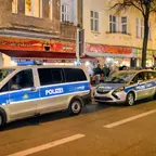 Zwei geparkte Polizeifahrzeuge vor einem beleuchteten Café mit mehreren Personen auf dem Bürgersteig