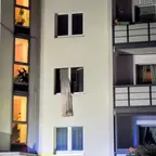 Fassade eines mehrstöckigen Wohnhauses bei Nacht mit offenem Fenster, aus dem ein Vorhang nach außen hängt, und beleuchteten Fenstern im Treppenhaus.